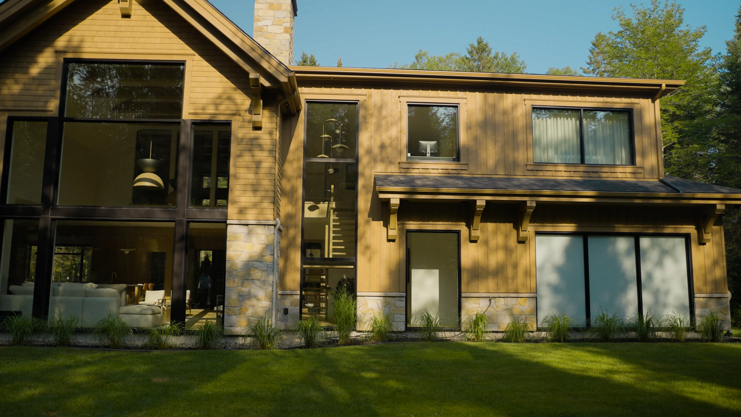 Façade d’une maison avec grandes fenêtres, porte patio et parement en bois et en pierre.